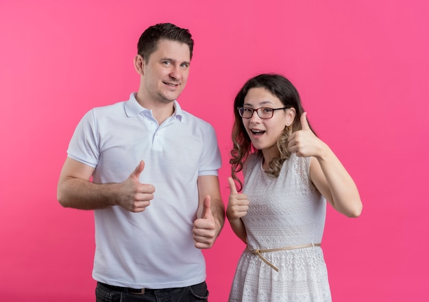 Junges Paar Mann und Frau in der Freizeitkleidung lächelnd fröhlich Daumen hoch stehend über rosa Wand