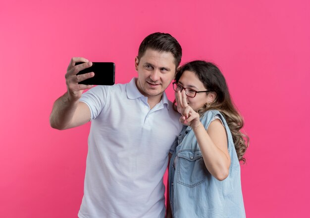 Junges Paar Mann und Frau in Freizeitkleidung unter Selfie lächelnd glücklich in der Liebe stehen zusammen über rosa Wand