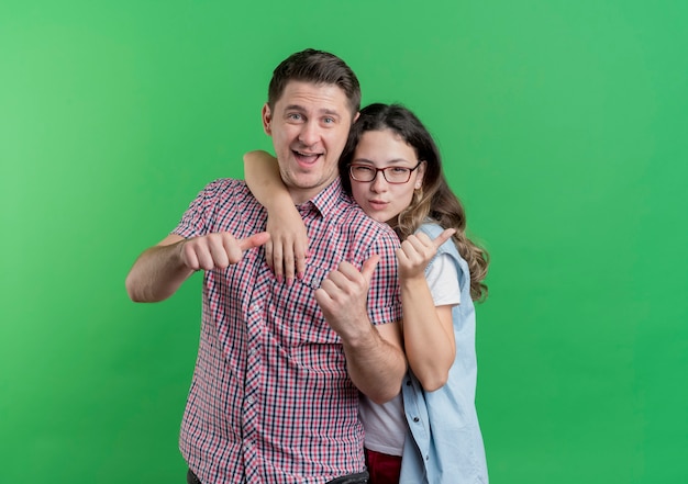 Junges Paar Mann und Frau in lässigen Kleidern lächelnd zeigt Daumen hoch über grüner Wand