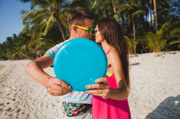 Junges Paar Mann und Frau spielen fliegende Scheibe am tropischen Strand, Sommerferien, Liebe, Romantik, fröhliche Stimmung, Lächeln, Spaß haben, Hipster-Outfit, Sonnenbrille, Jeansshorts, sonnig, positive Stimmung