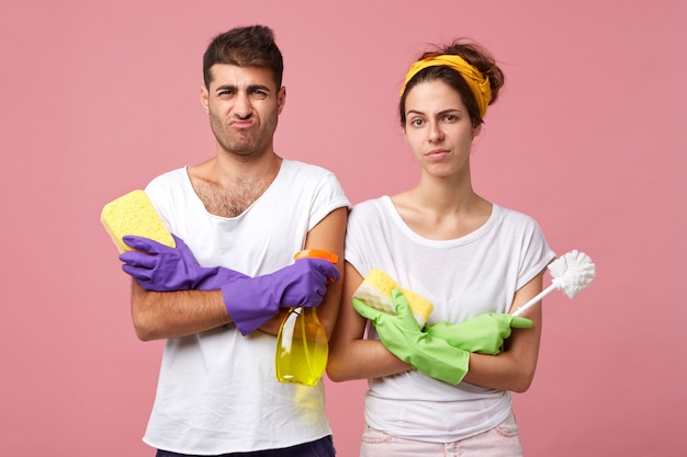 Junges Paar mit stirnrunzelnden Gesichtern, die ihre Hände gekreuzt halten und Bürste, Waschmittel und Mops tragen, tragen Schutzhandschuhe und weiße lässige T-Shirts, die schlechte Laune haben und nicht bereit sind, das Haus zu putzen