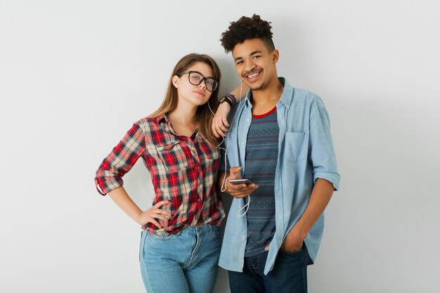 Junges Paar, schwarzer Hipster-Mann, kaukasisches Mädchen auf lokalem weißem Hintergrund, stilvolles Outfit, Freunde, die zusammen Spaß haben, Musik auf Kopfhörern hören, Smartphone halten, lächelnd, glücklich