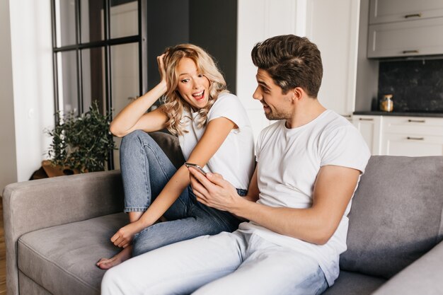 Junges Paar sitzt auf der Couch. Aufgeregter Mann zeigt seiner Freundin Goon News auf seinem Handy. Überraschtes Mädchen, das auf den Telefonbildschirm schaut und glücklich lächelt.