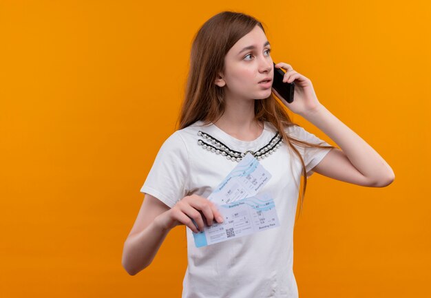 Junges reisendes Mädchen, das am Telefon spricht und Flugtickets hält, die rechte Seite auf isolierter orange Wand mit Kopienraum betrachten