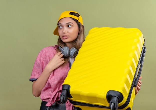 Junges reisendes Mädchen, das rosa Hemd in der Kappe mit Kopfhörern um Hals hält, der Koffer hält, der verwirrt schaut