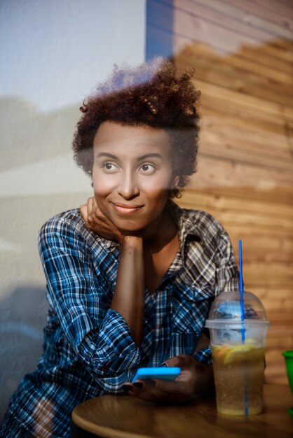 Junges schönes afrikanisches Mädchen lächelnd, Telefon haltend, im Café sitzend. Von außen geschossen.