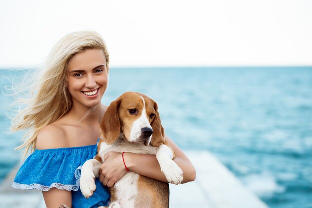 Junges schönes blondes Mädchen, das mit Beagle-Hund am Meer spielt.