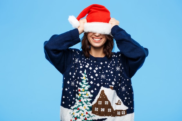 Junges schönes brünettes Mädchen im gestrickten Pullover und in der Weihnachtsmütze, die über blaue Wand lächeln