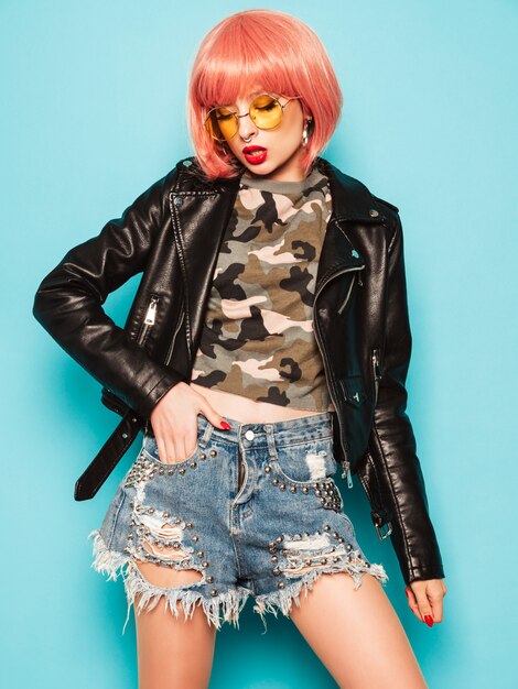 Junges schönes hippes böses Mädchen in der schwarzen Lederjacke und im Ohrring in der Nase. Sexy sorglose Frau, die im Studio in der rosa Perücke nahe blauer Wand aufwirft. Selbstbewusstes Modell in der Sonnenbrille