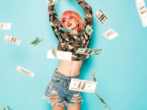 Junges schönes hipster böses mädchen in trendigen roten sommerkleidern und ohrring in der nase. sexy sorglose frau, die im studio in der rosa perücke nahe blauer wand sitzt. modell, das gelddollar-banknoten herauswirft