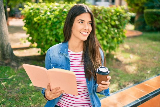 Junges schönes hispanisches Mädchen, das glückliches Lesebuch lächelt und Kaffee trinkt, der am Park sitzt