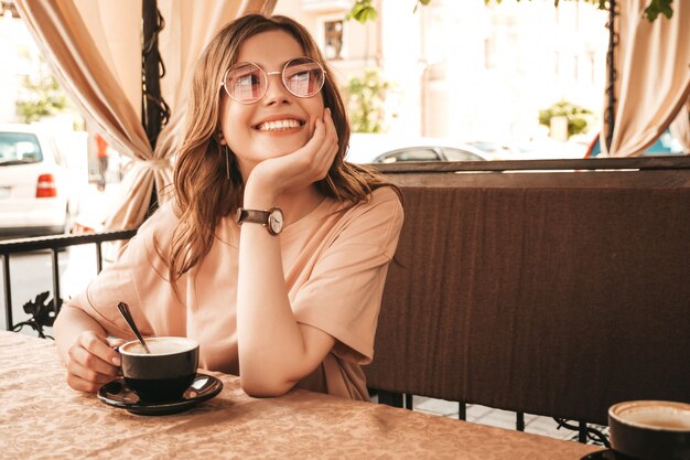 Junges schönes lächelndes Hipster-Mädchen in den trendigen Sommerkleidern. Sorglose Frau, die im Veranda-Terrassencafé sitzt und Kaffee trinkt. Positives Modell, das Spaß und Träume hat