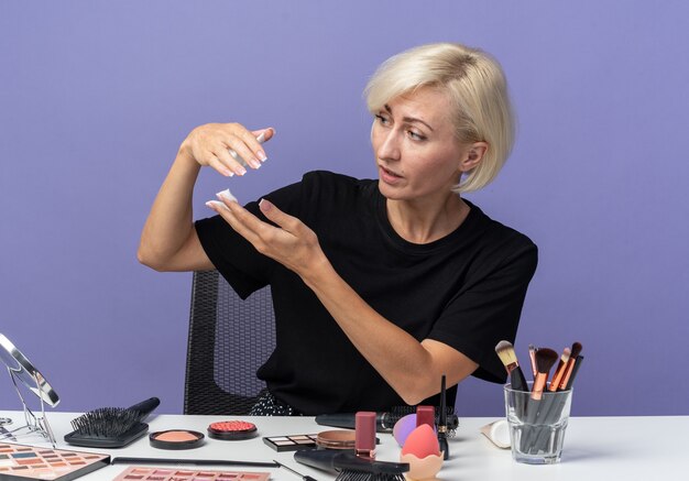 Junges schönes Mädchen auf der Suche nach Seite sitzt am Tisch mit Make-up-Tools, die Haarcreme isoliert auf blauer Wand halten