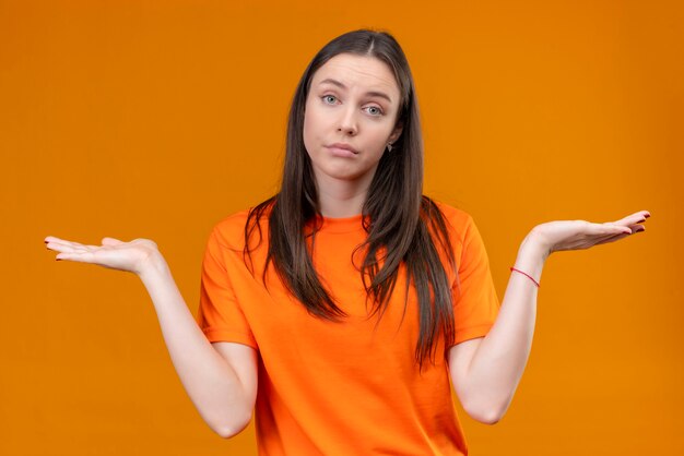 Junges schönes Mädchen, das ahnungsloses orange T-Shirt und verwirrte achselzuckende Schultern trägt, die Hände verbreiten, die keine Antwort haben, die über lokalisiertem orangefarbenem Hintergrund steht