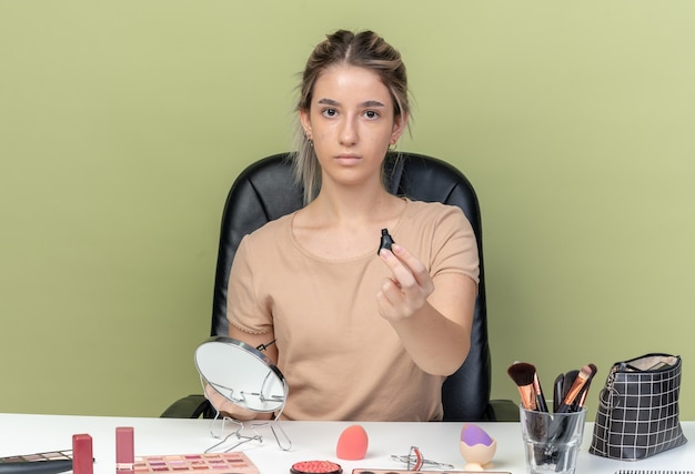 Junges schönes Mädchen, das am Schreibtisch mit Make-up-Tools sitzt und Wimperntusche in die Kamera hält, isoliert auf olivgrüner Wand?