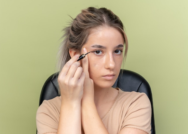 Junges schönes mädchen, das am tisch mit make-up-tools sitzt, zeichnet pfeil mit eyeliner isoliert auf olivgrüner wand