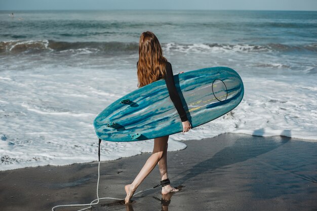 junges schönes Mädchen, das auf dem Strand mit einem Surfbrett, Frauensurfer, Meereswogen aufwirft
