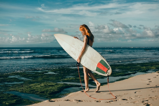 junges schönes Mädchen, das auf dem Strand mit einem Surfbrett, Frauensurfer, Meereswogen aufwirft