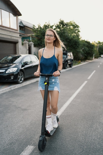 Junges schönes Mädchen, das einen Elektroroller im Sommer auf der Straße reitet