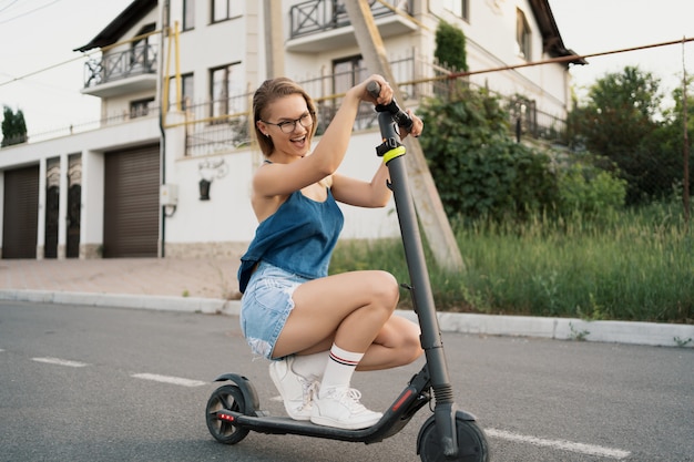 Junges schönes Mädchen, das einen Elektroroller im Sommer auf der Straße reitet