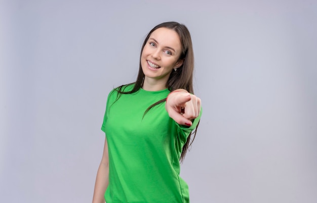 Junges schönes Mädchen, das grünes T-Shirt glücklich und positiv lächelnd fröhlich zeigt auf Kamera steht über lokalisiertem weißem Hintergrund trägt