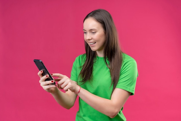 Junges schönes Mädchen, das grünes T-Shirt hält, das Smartphone hält, das mit jemandem glücklich und positiv lächelnd steht über lokalisiertem rosa Hintergrund steht