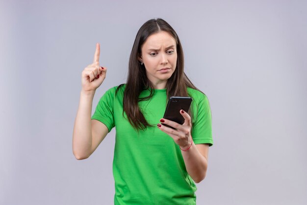 Junges schönes Mädchen, das grünes T-Shirt hält, das Smartphone zeigt Finger zeigt, der sich daran erinnert, über isoliertem weißem Hintergrund zu stehen