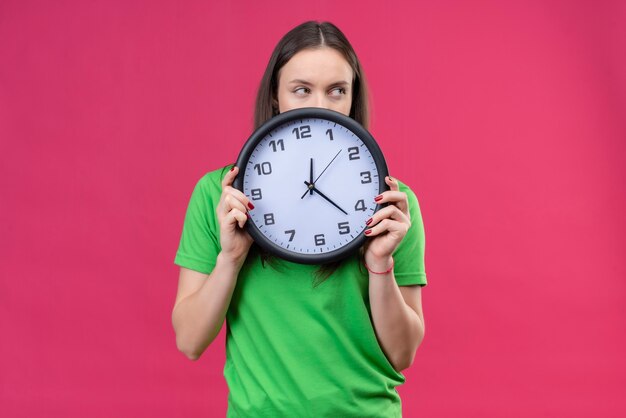 Junges schönes Mädchen, das grünes T-Shirt hält Uhr hält, das über es steht, das über lokalisiertem rosa Hintergrund steht