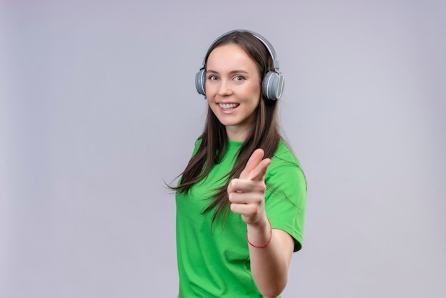 Junges schönes Mädchen, das grünes T-Shirt mit Kopfhörern trägt, die positiv und glücklich lächeln, zeigen auf Kamera, die über lokalisiertem weißem Hintergrund steht