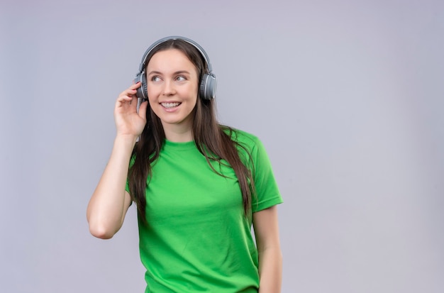 Junges schönes Mädchen, das grünes T-Shirt mit Kopfhörern trägt und ihre Lieblingsmusik genießt, die fröhlich über lokalisiertem weißem Hintergrund stehend lächelt