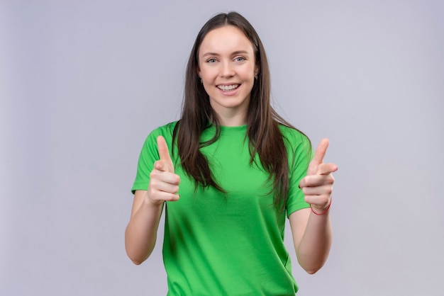 Junges schönes Mädchen, das grünes T-Shirt trägt, das fröhlich auf Kamera mit beiden Händen zeigt, die über lokalisiertem weißem Hintergrund stehen