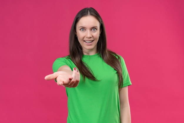 Junges schönes Mädchen, das grünes T-Shirt trägt, das Kamera mit glücklichem Gesicht macht, kommt hierher Geste mit Hand, die über lokalisiertem rosa Hintergrund steht