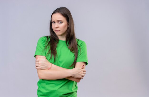 Junges schönes Mädchen, das grünes T-Shirt trägt, das mit verschränkten Armen steht und Kamera unzufrieden steht, die über lokalisiertem weißem Hintergrund steht