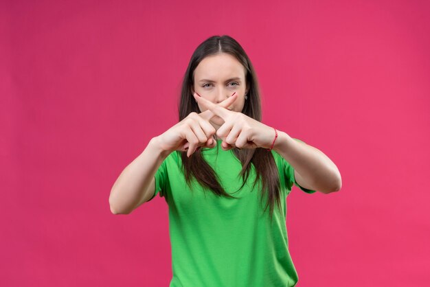 Junges schönes Mädchen, das grünes T-Shirt trägt, das Verteidigungsgeste macht, die Zeigefinger kreuzt, die mit stirnrunzelndem Gesicht stehen, das über lokalisiertem rosa Hintergrund steht