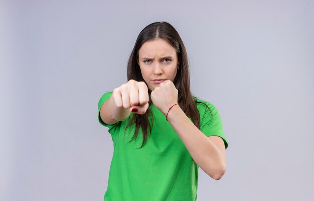 Junges schönes Mädchen, das grünes T-Shirt trägt, das wie eine Boxer-geballte Faust zur Kamera aufwirft, die mit stirnrunzelndem Gesicht steht, das über lokalisiertem weißem Hintergrund steht