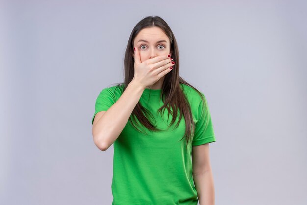 Junges schönes Mädchen, das grünes T-Shirt trägt, schockierte das Abdecken des Mundes mit der Hand, die über lokalisiertem weißem Hintergrund steht