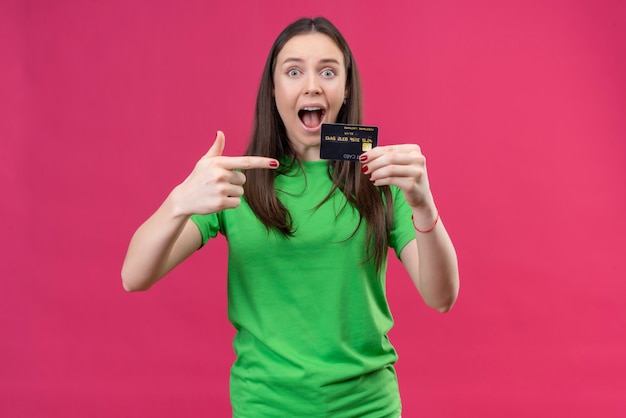 Junges schönes Mädchen, das grünes T-Shirt trägt, verließ und glücklich hält Kreditkarte, die mit dem Finger darauf zeigt, der über lokalisiertem rosa Hintergrund steht