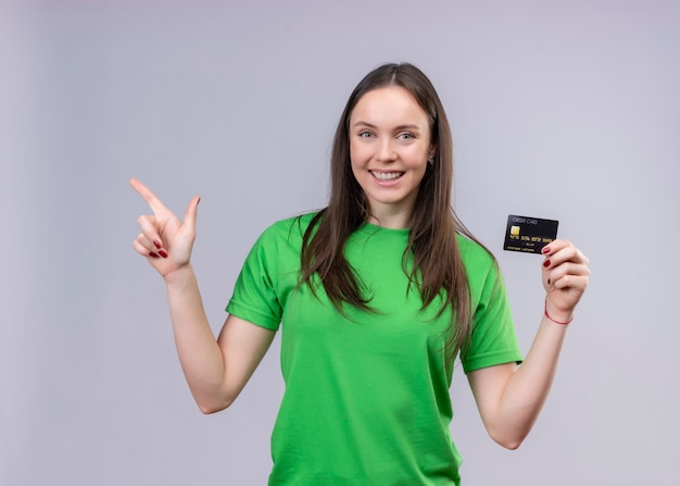 Junges schönes Mädchen, das grünes T-Shirt trägt, verließ und glücklich hält Kreditkarte, die mit Finger zur Seite steht, die über lokalisiertem weißem Hintergrund steht