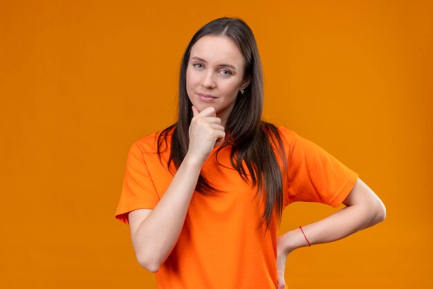 Junges schönes Mädchen, das orange T-Shirt betrachtet Kamera mit Hand auf Kinn denkt, das über lokalisiertem orange Hintergrund steht