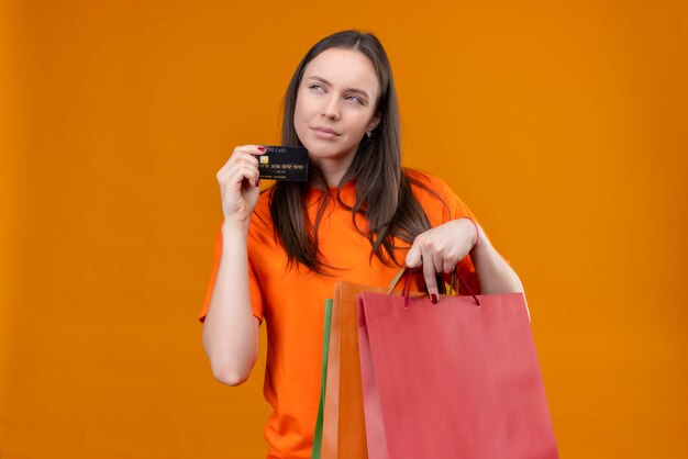 Junges schönes Mädchen, das orange T-Shirt hält, das Papierpaket und Kreditkarte hält, die schlau beiseite stehend über lokalisiertem orange Hintergrund stehen