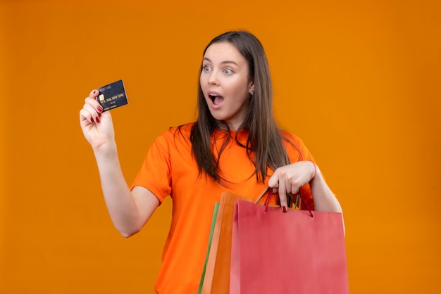 Junges schönes Mädchen, das orange T-Shirt hält Papierpaket und Kreditkarte lächelnd fröhlich überrascht und verlassen steht über isoliertem orange Hintergrund