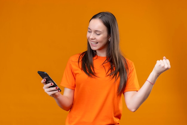 Junges schönes Mädchen, das orange T-Shirt hält Smartphone hält, das es glücklich und positiv lächelnd über lokalisiertem orange Hintergrund betrachtet