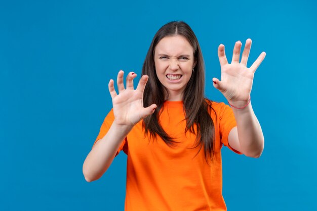 Junges schönes Mädchen, das orange T-Shirt knurrt, das wie Tier macht, das Katzenkrallen gestikuliert, das über lokalisiertem blauem Hintergrund steht