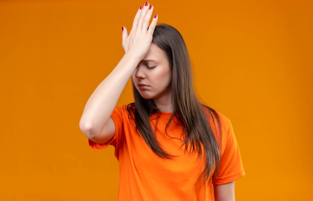 Junges schönes mädchen, das orange t-shirt trägt, das mit hand auf kopf für fehler steht, der mit traurigem ausdruck auf gesicht über lokalisiertem orange hintergrund unglücklich schaut