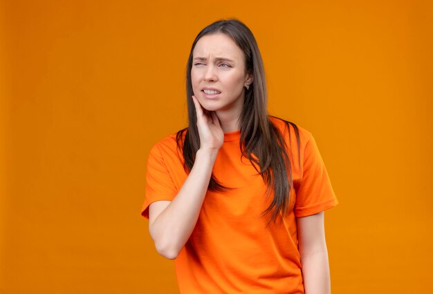 Junges schönes Mädchen, das orange T-Shirt trägt, das unwohl schaut, das ihre Wange berührt, die Zahnschmerzen fühlt, die über lokalisiertem orange Hintergrund stehen