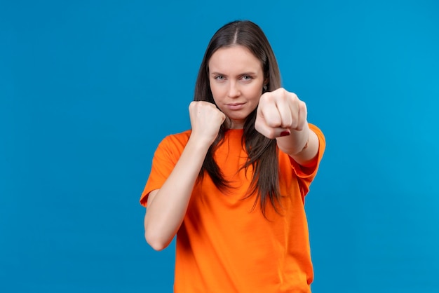 Junges schönes Mädchen, das orange T-Shirt trägt, das wie ein Boxer aufwirft, der Faust zur Kamera zeigt, die zuversichtlich steht, über lokalisiertem blauem Hintergrund zu stehen