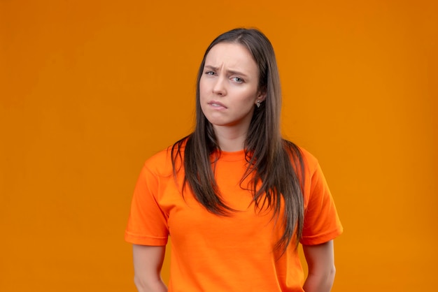 Junges schönes Mädchen, das orange T-Shirt trägt Kamera betrachtet unzufrieden mit traurigem Ausdruck auf Gesicht, das über lokalisiertem orange Hintergrund steht
