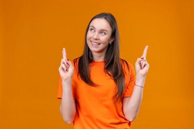 Junges schönes Mädchen, das orange T-Shirt trägt, wünschenswert wünschenswert mit kreuzenden Fingern lächelnd fröhlich stehend über isoliertem orange Hintergrund