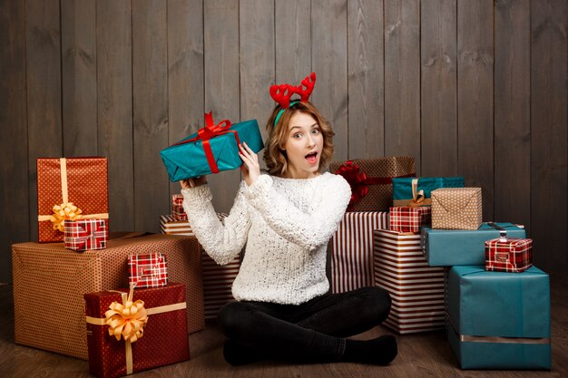 Junges schönes Mädchen, das unter Weihnachtsgeschenken über Holzwand sitzt