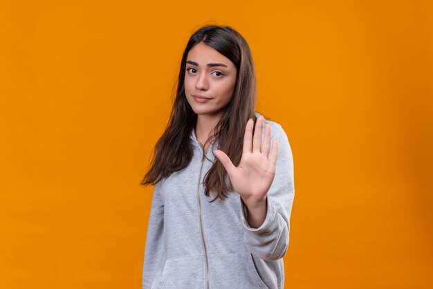 Junges schönes Mädchen im grauen Kapuzenpulli, das den müden Anblick der Kamera auf Gesicht betrachtet und Stoppgeste macht, die über orange Hintergrund steht